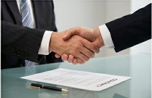 Two individuals in business suits shaking hands above a desk with a document labeled "CONTRACT" and a pen lying below them.